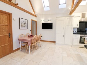 A dining area with a table set for four, a television mounted on the wall, and a kitchen with white cabinets and a stove. - Sutton Newbold Cottage (Trimdon Station)
