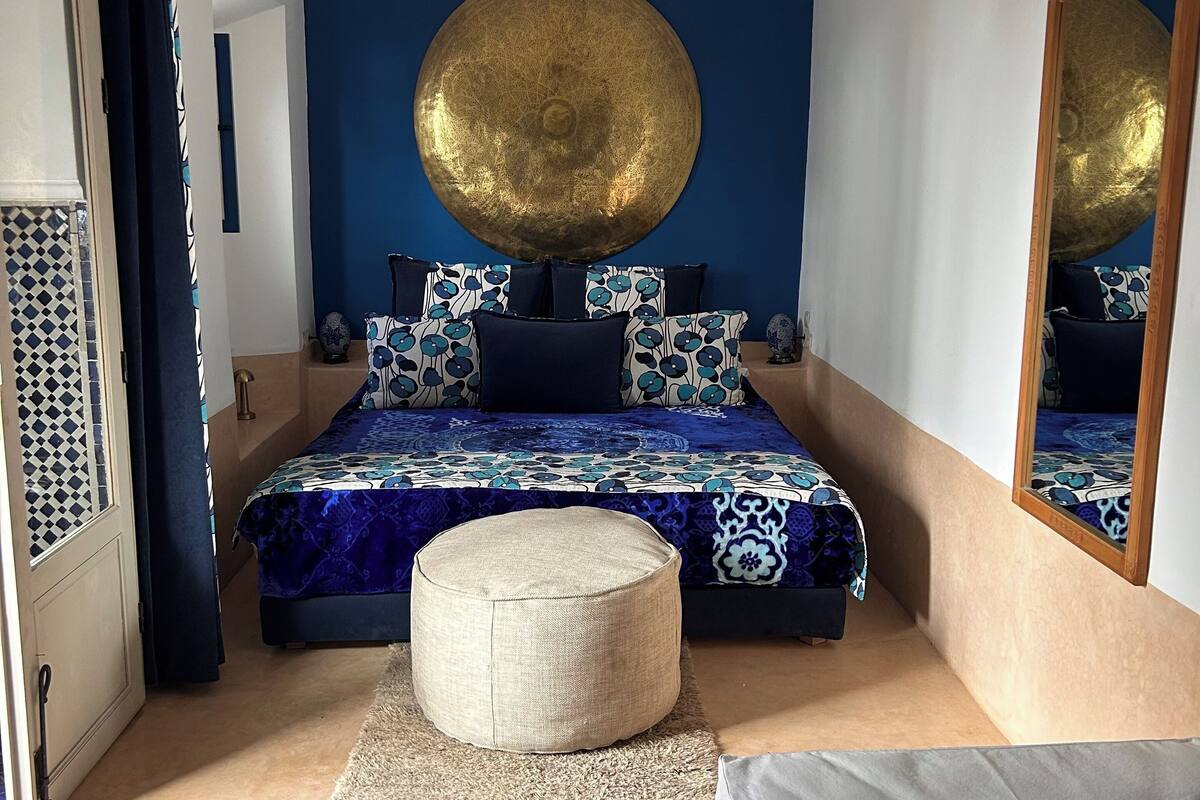 A bedroom with a large bed, a round decorative item on the wall, a round ottoman, and a mirror.