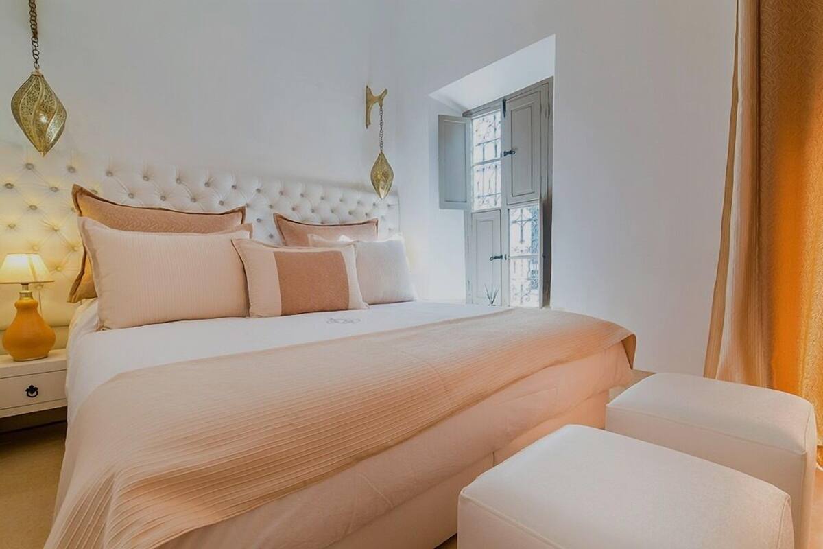 A bedroom with a tufted headboard, a large bed with multiple pillows, a nightstand with a lamp, and a wardrobe with a window.