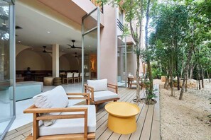Une terrasse extĂ©rieure avec des meubles en bois et une table jaune, entourĂ©e dâarbres et dâun bĂątiment dotĂ© de grandes fenĂȘtres.