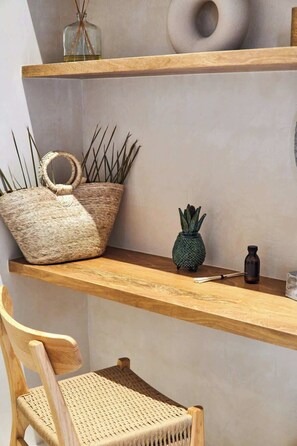 Una sedia di legno con seduta intrecciata, un cesto di vimini con manici, una piccola pianta in vaso e una bottiglia nera su uno scaffale di legno.