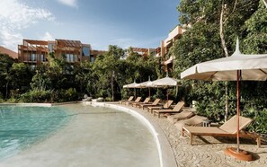 Un espace aménagé au bord de la piscine, avec des chaises longues et des parasols, entouré de verdure et de bùtiments.