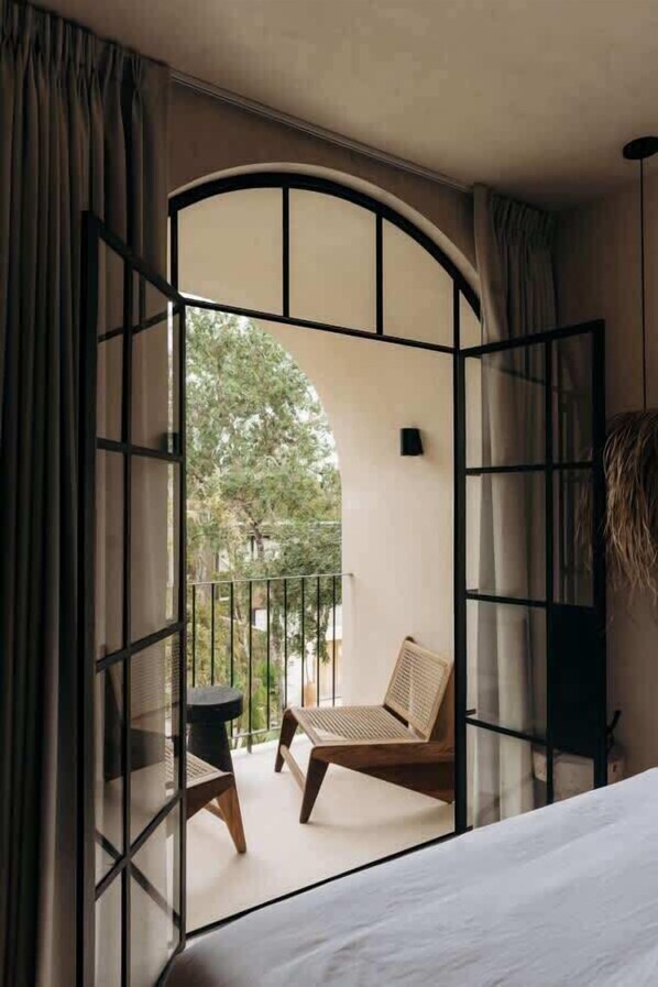 Una habitación con una cama, una mesita y una silla. Tiene un balcón con vistas a árboles.