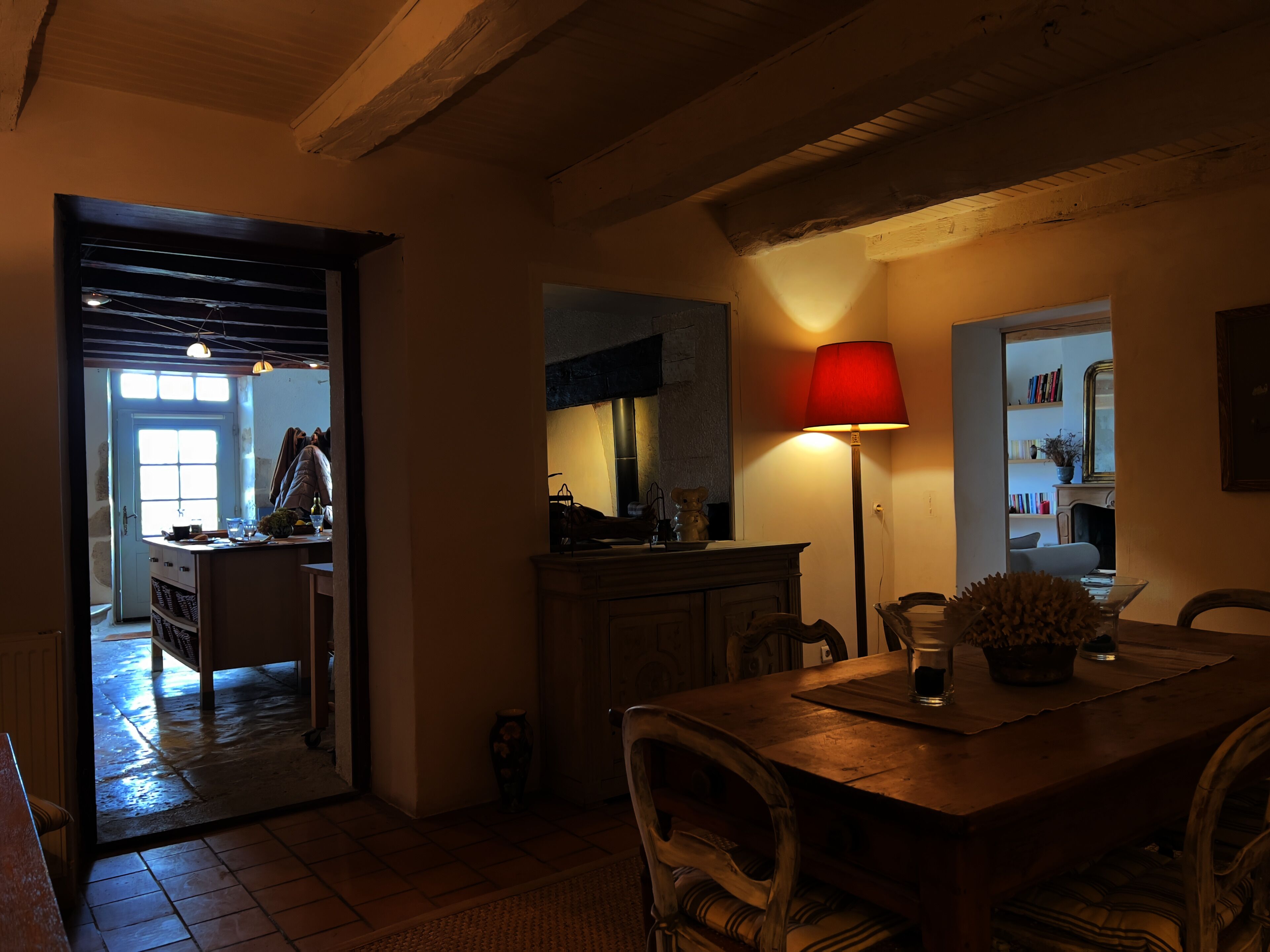 Un coin repas accueillant avec une table en bois, des chaises et une lampe rouge. On aperçoit une cuisine à l’arrière-plan, avec une fenêtre et une porte donnant sur l’extérieur.