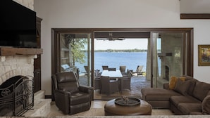 Amplia sala de estar con chimenea de piedra, sillón de cuero y vista a un lago a través de puertas de vidrio grandes.