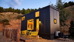 Een moderne, compacte cabin met een houten terras en gele stoelen, omgeven door een rustieke omheining en een natuurlijk landschap.
