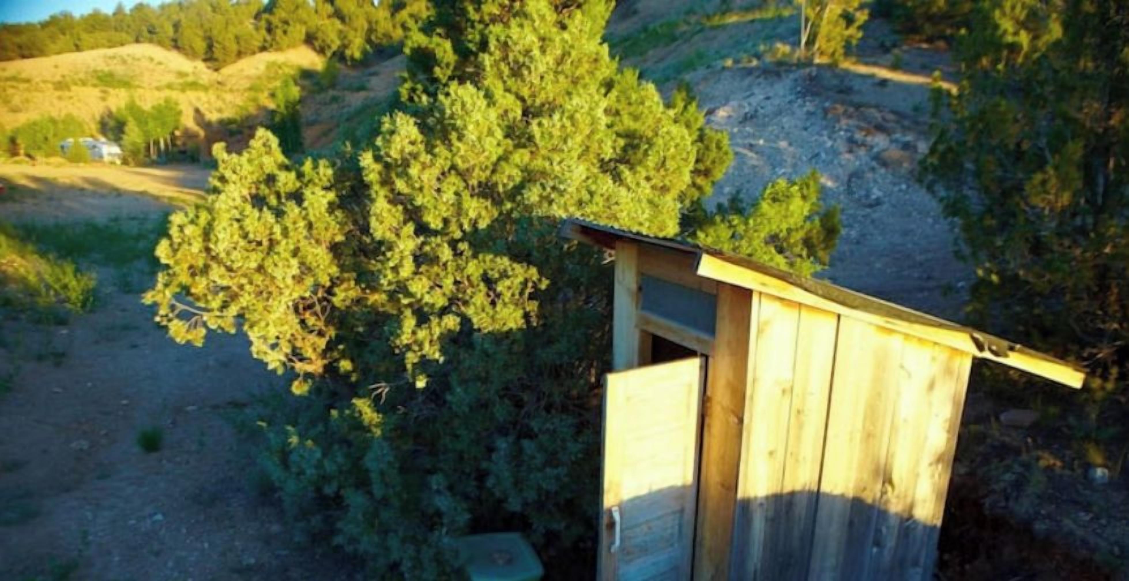 A wooden outhouse with a slanted roof, surrounded by greenery and hills.
