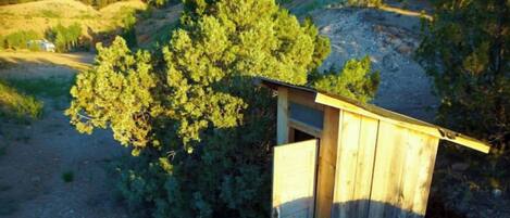 A wooden outhouse with a slanted roof, surrounded by greenery and hills.