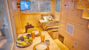 Een klein houten vakantiehuisje met een bed, een eettafel met fruit en glazen, en een televisie aan de muur.
