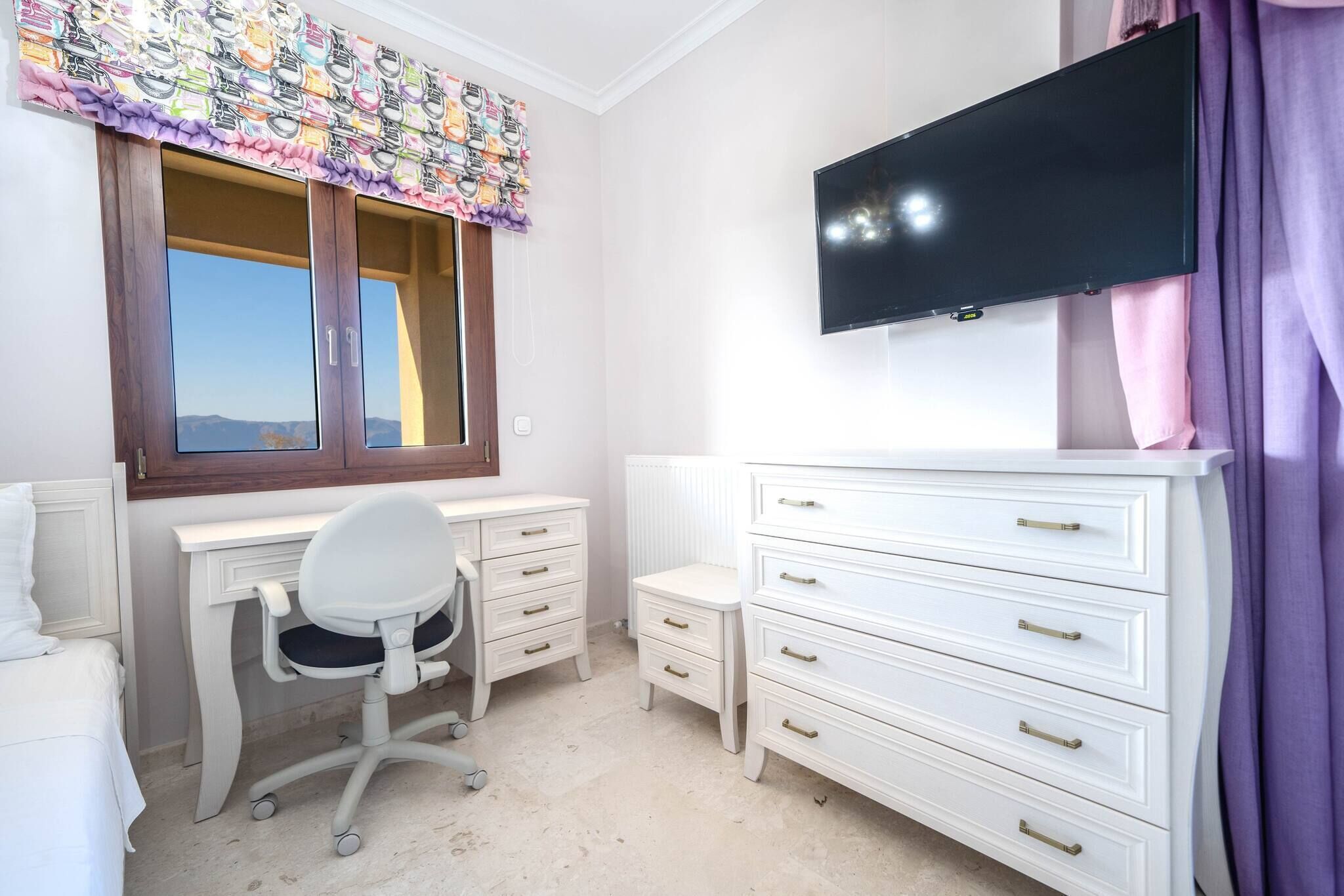 Quarto com cama, escrivaninha, cadeira, televisão e cômoda.