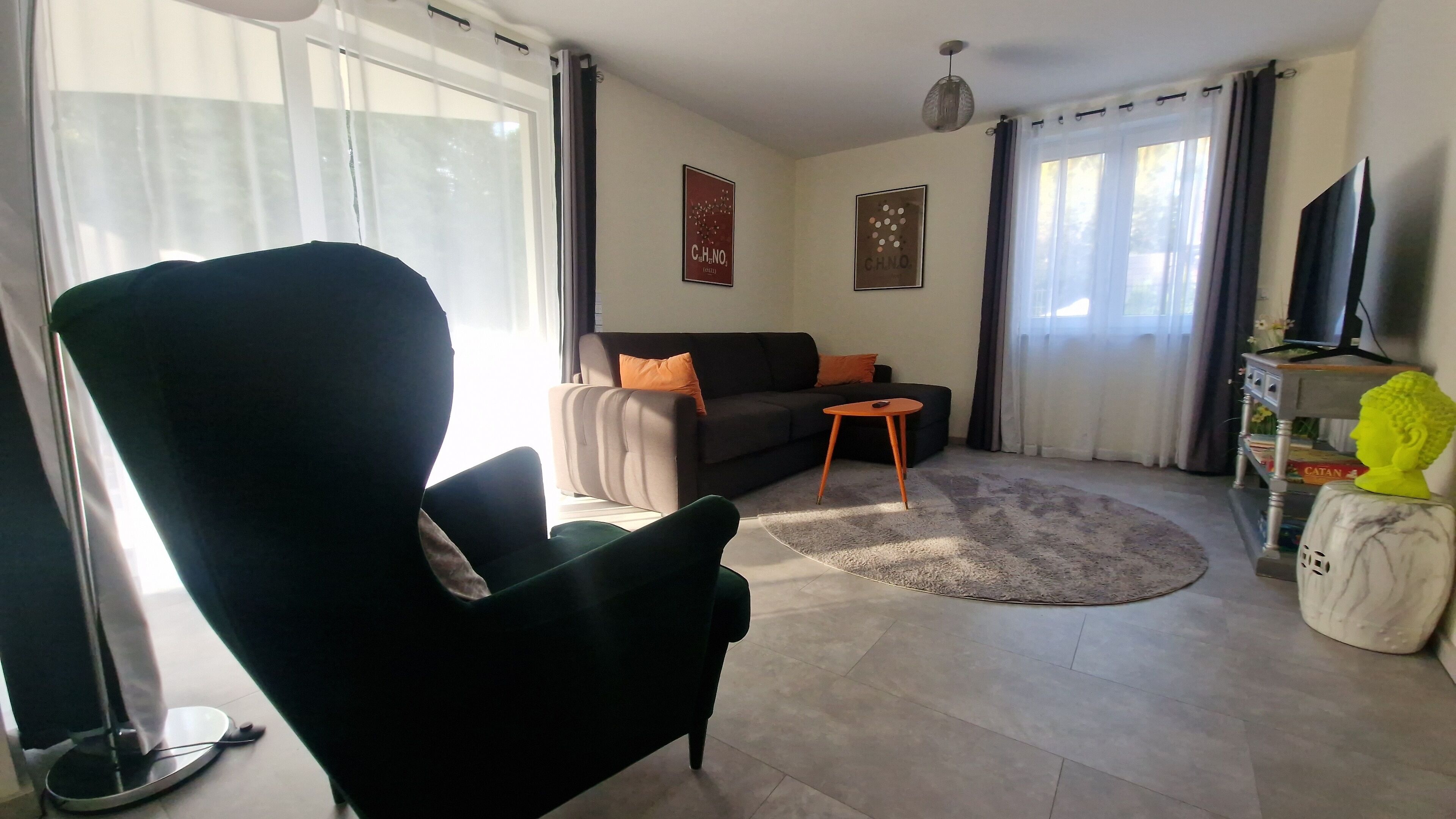Een woonkamer met een grote zwarte fauteuil, een grijze bank met oranje kussens, een klein rond tafeltje en een televisie op een dressoir.