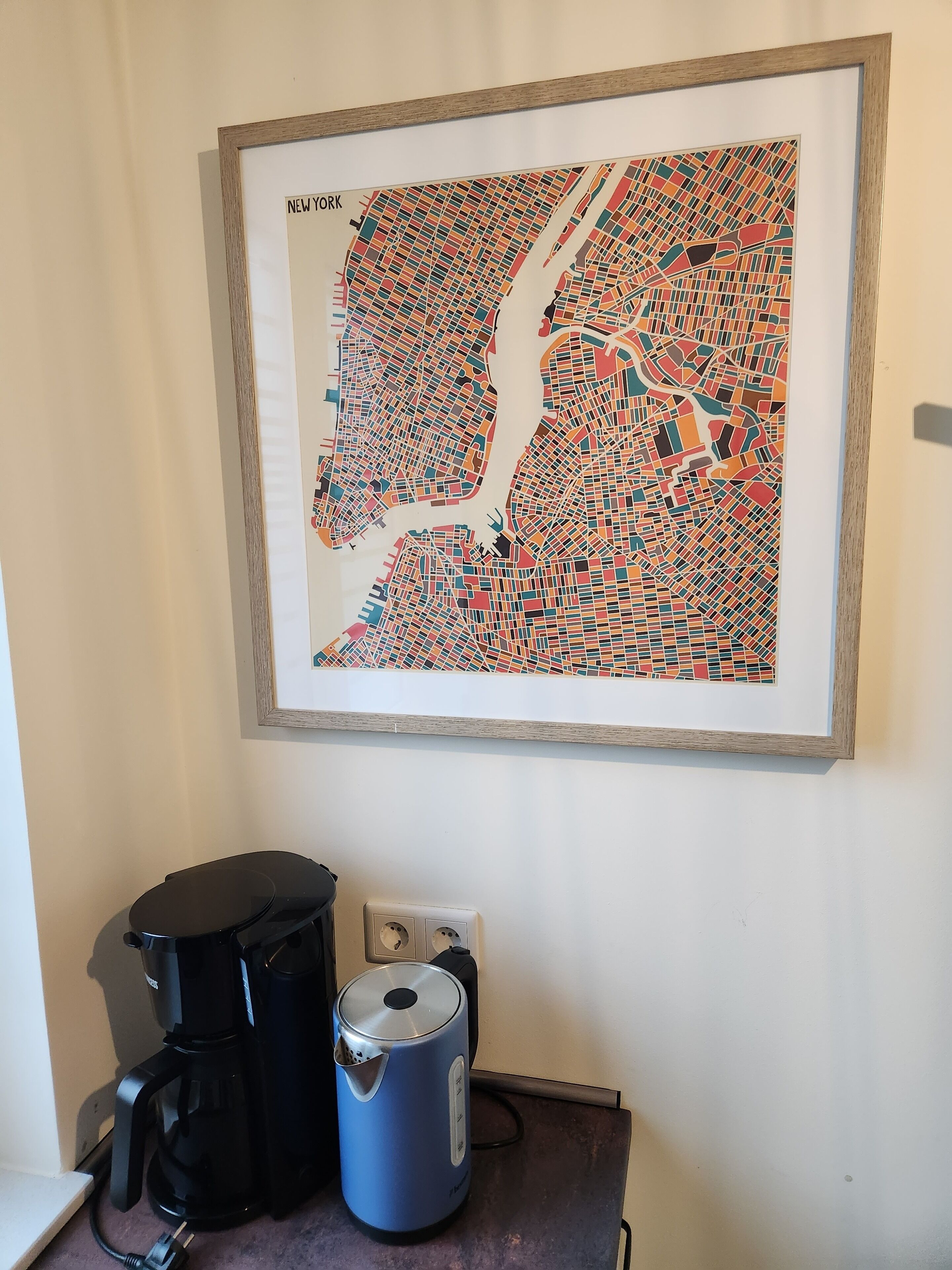 Un mapa enmarcado de Nueva York en una pared, una cafetera de color negro y una tetera azul sobre una encimera.