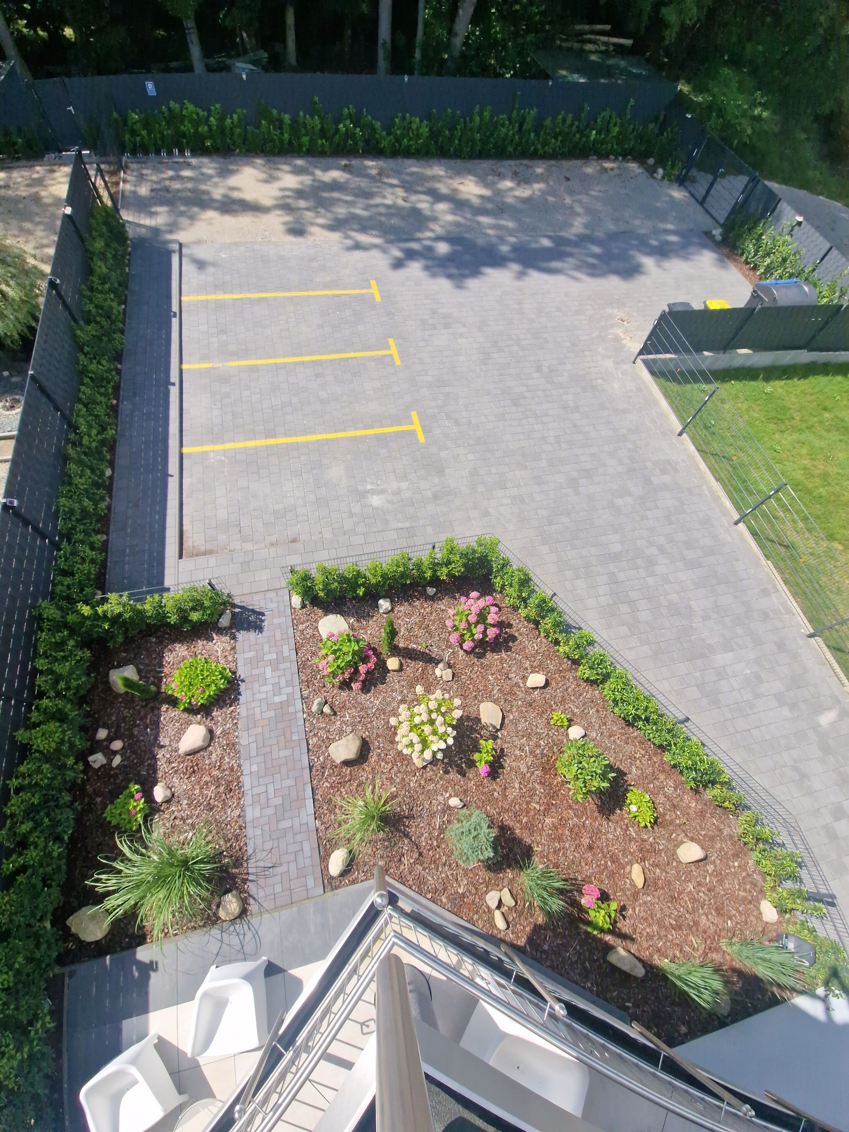 Ein Parkplatz mit markierten Stellplätzen, ein angelegter Garten mit verschiedenen Pflanzen und Steinen sowie ein Metallgeländer entlang des Randes.