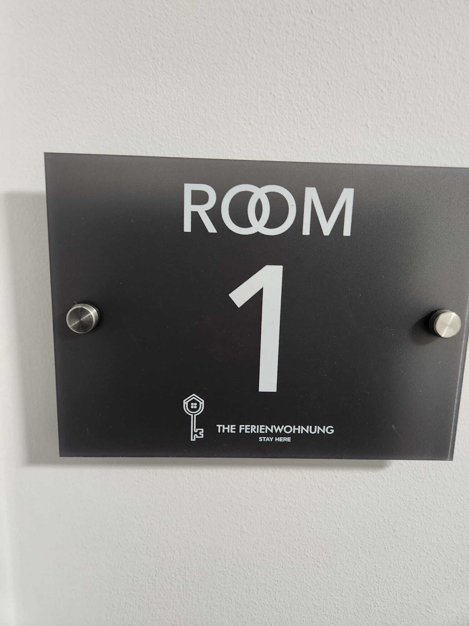 Un senyal negre amb text blanc que indica “HABITACIÓ 1” i el logotip de “THE FERIENWOHNUNG” amb la frase “QUANTAL’ESTADA”. 