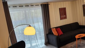 Un salon avec un canapé noir, un fauteuil noir, une table basse ronde, une suspension jaune et une fenêtre avec des rideaux.