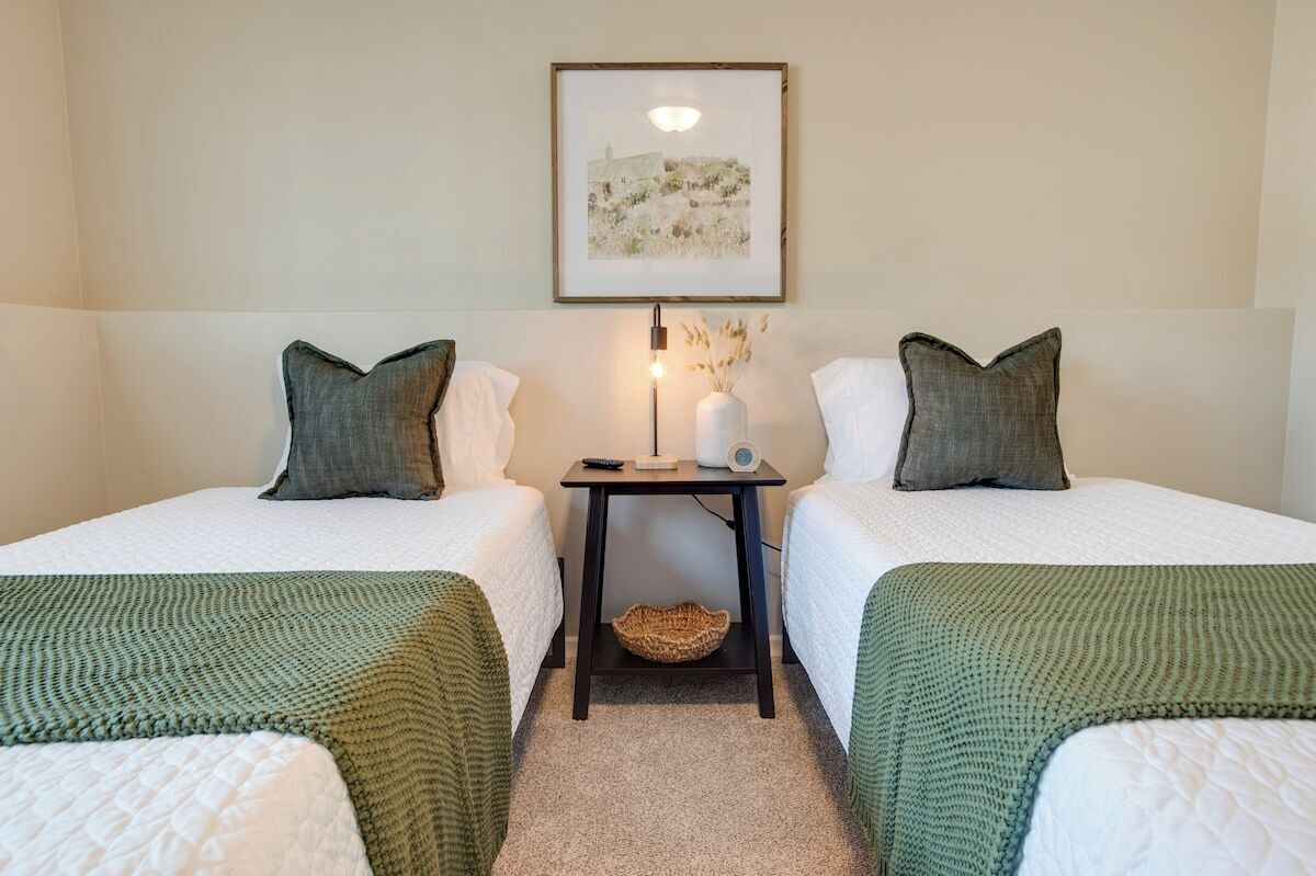 A hotel room with two single beds, a small table with a lamp, a framed picture on the wall, and a basket on the floor.