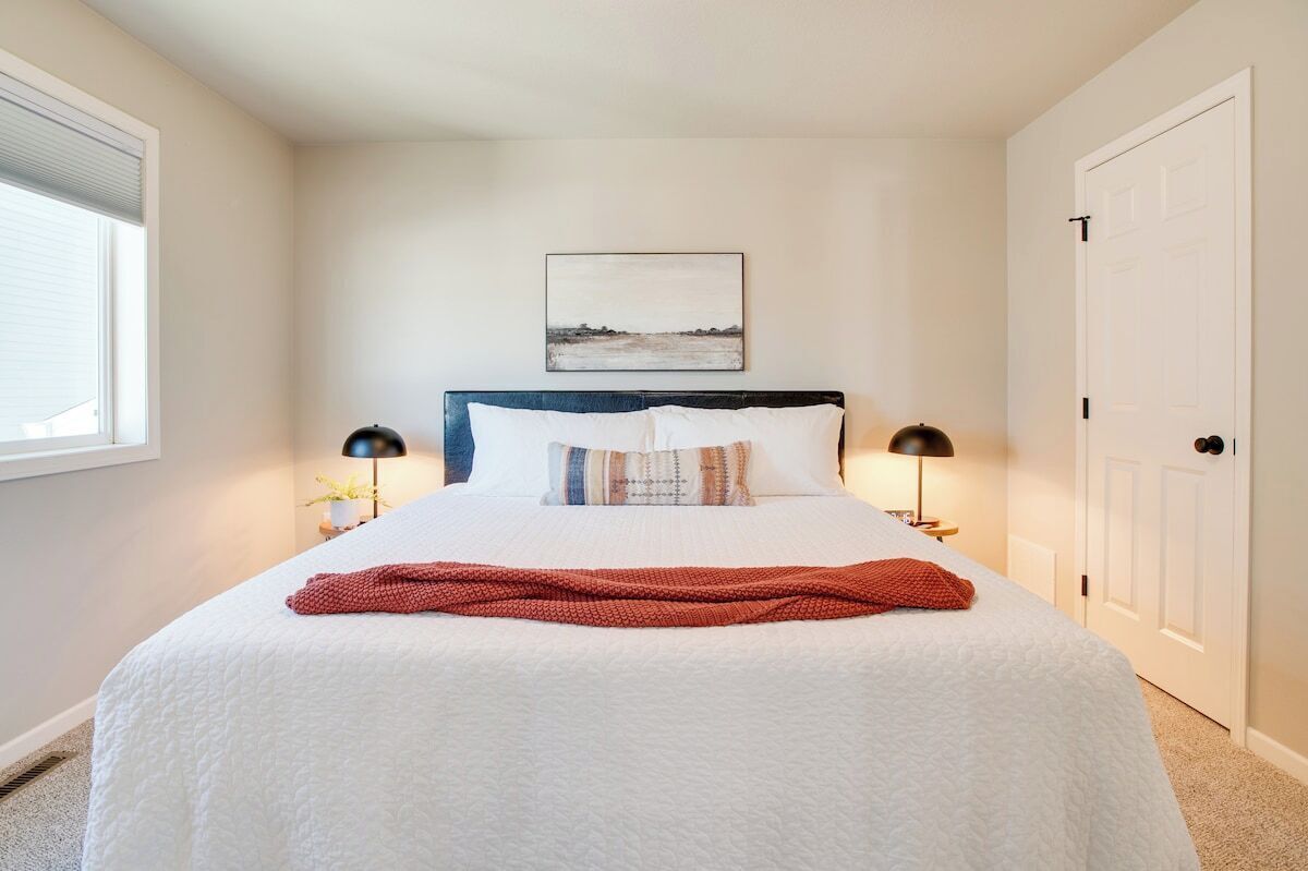 Une chambre à coucher bien rangée, avec un grand lit, deux lampes de chevet, un tableau encadré au mur et une porte sur le côté droit.