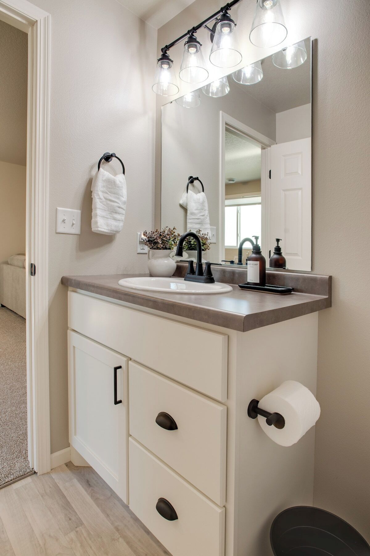 Une salle de bain avec un meuble-lavabo blanc, un miroir avec éclairage intégré, un porte-serviettes, un lavabo, un porte-rouleau de papier toilette et une petite plante en pot.