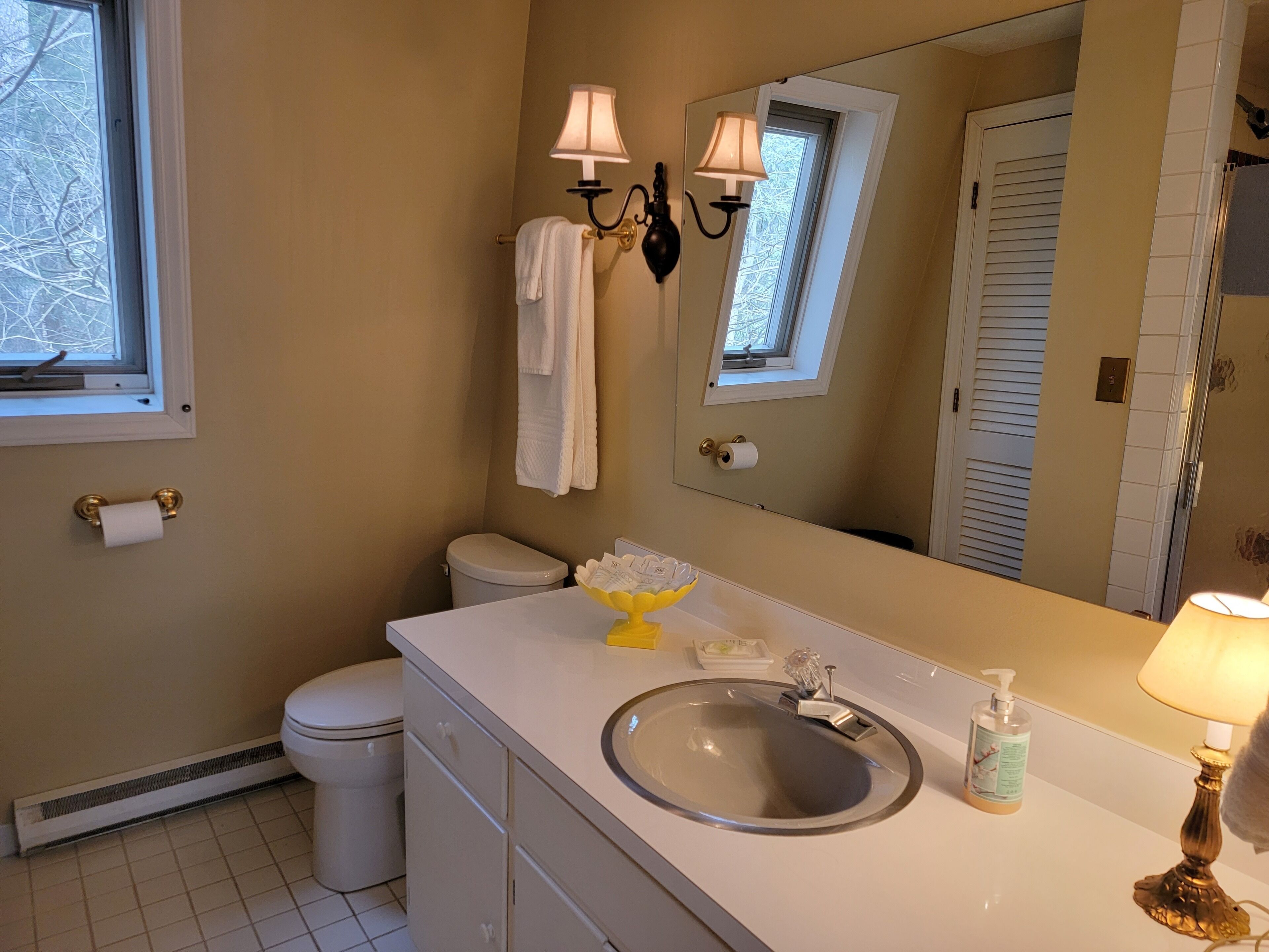 A bathroom with a toilet, sink, and a mirror. There are towels hanging on the wall, a window, and a door.