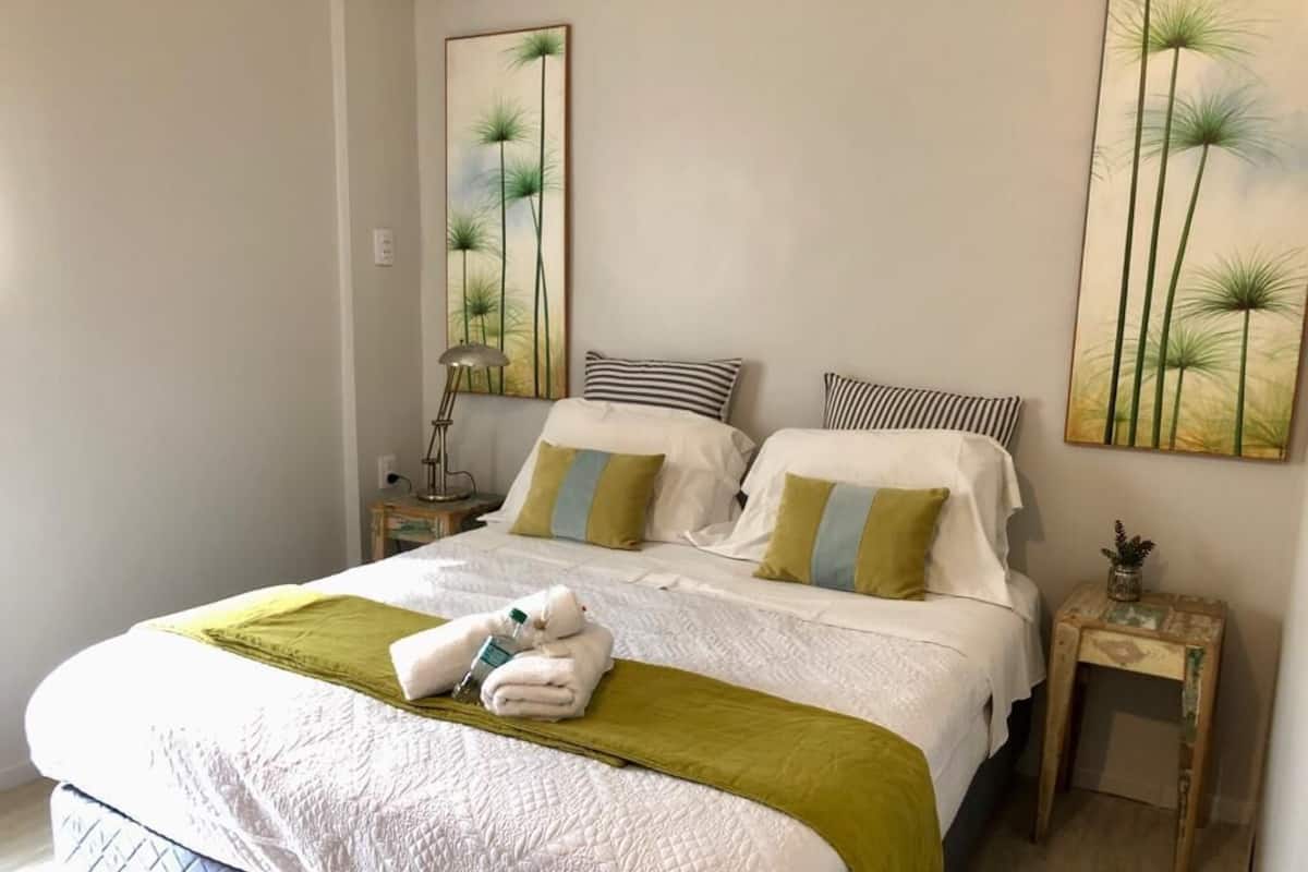 Una cama bien hecha con edredón blanco y manta verde, dos mesitas de noche con lámparas, y dos cuadros en las paredes.