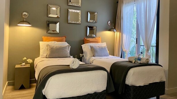 Ein Doppelzimmer mit zwei Betten, jeweils mit Kissen und Decke. An der Wand befinden sich Spiegel und ein kleiner Tisch mit einem Blumenstrauß.