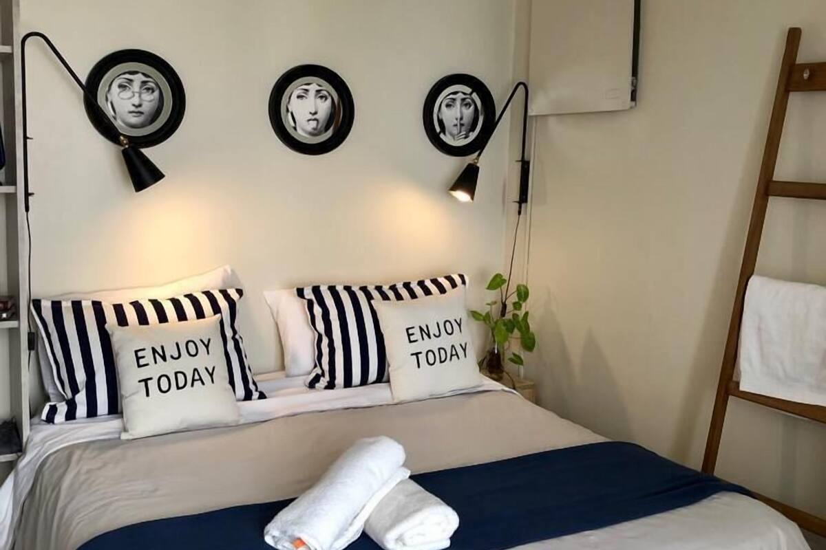 Una cama bien tendida con almohadillas a rayas y una manta azul, una escalera de madera y marcos de cuadros en la pared.