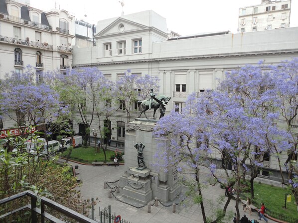 A courtyard with a statue, surrounded by buildings and trees with purple flowers.