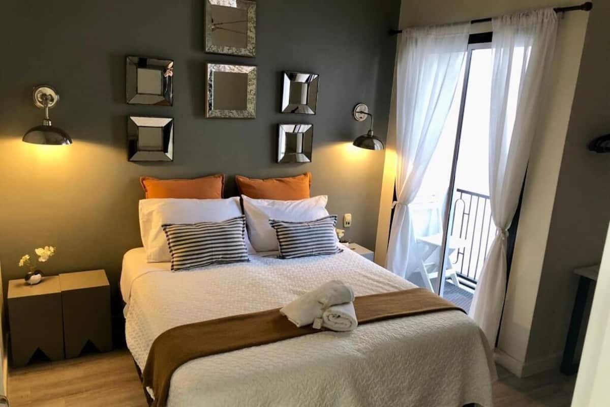 Una cama bien hecha con edredón blanco, dos almohadas y una manta marrón. Hay dos mesitas de noche con lámparas, un espejo en la pared y una ventana con cortinas.