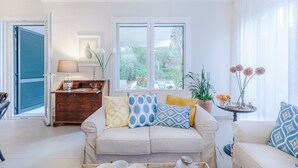 A living room with a beige sofa, patterned cushions, a wooden side table, and a view through large windows.