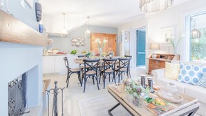 A spacious living area with a dining table, chairs, and a sofa. There is a kitchen area with a dining table and chairs, and a window with blue shutters.