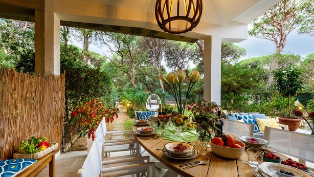 An outdoor dining area with a wooden table set for a meal, surrounded by lush greenery and trees.