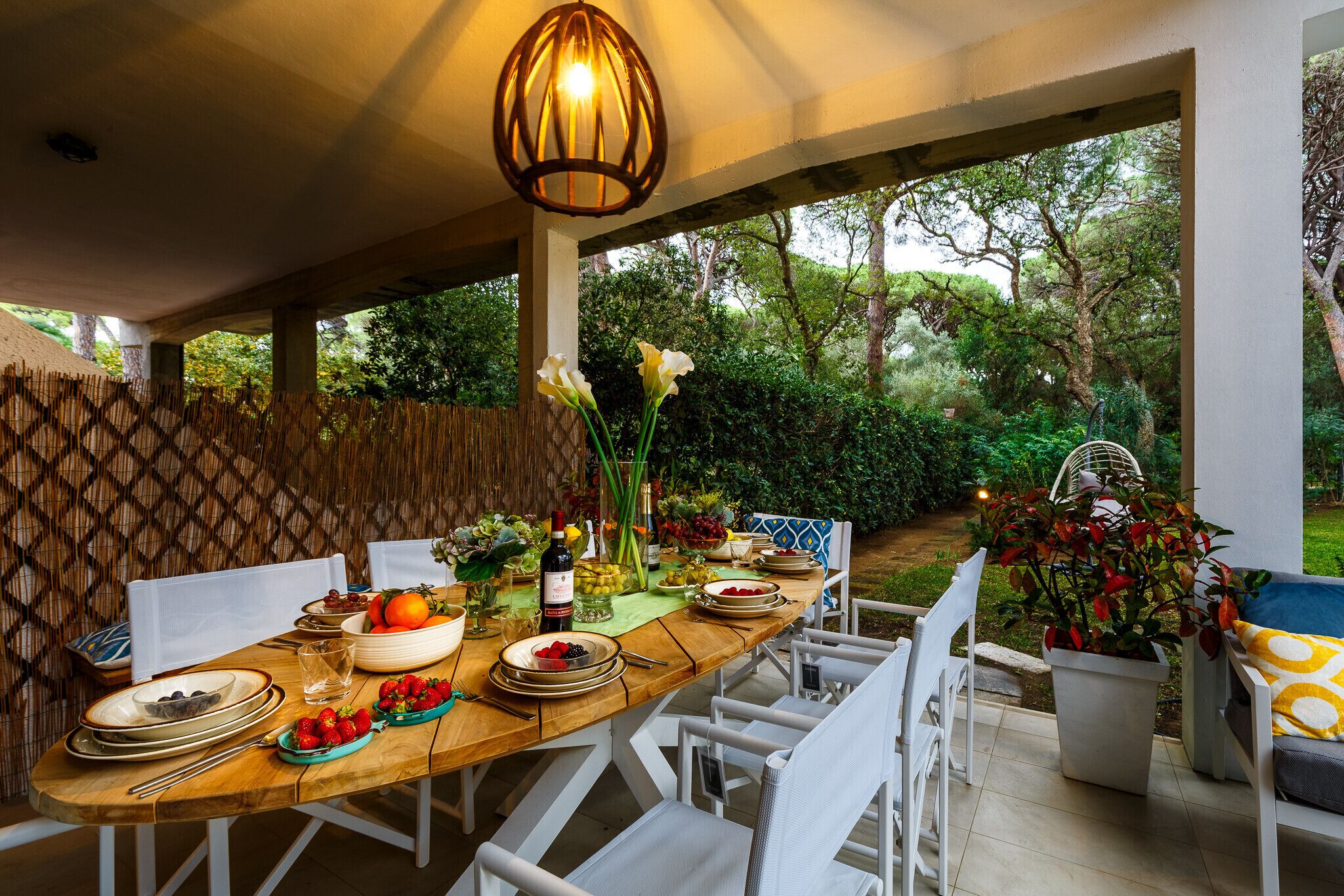 Un área de comedor al aire libre con una mesa de madera dispuesta para una comida, rodeada de sillas blancas y un colgante sobre ella.