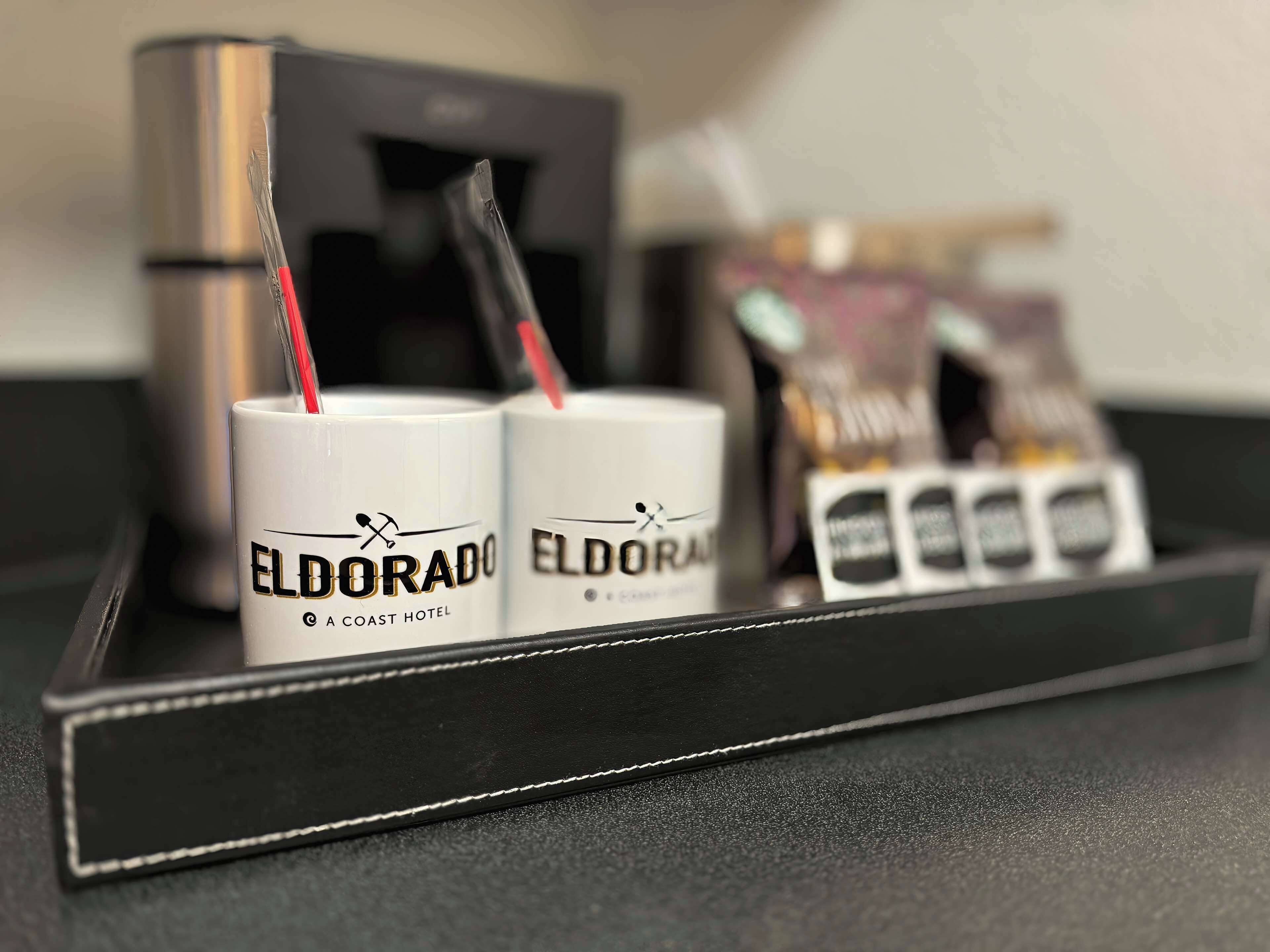 Twee witte mokken met 'Eldorado'-branding en een dienblad met een zwart object en een zak zakdoekjes.