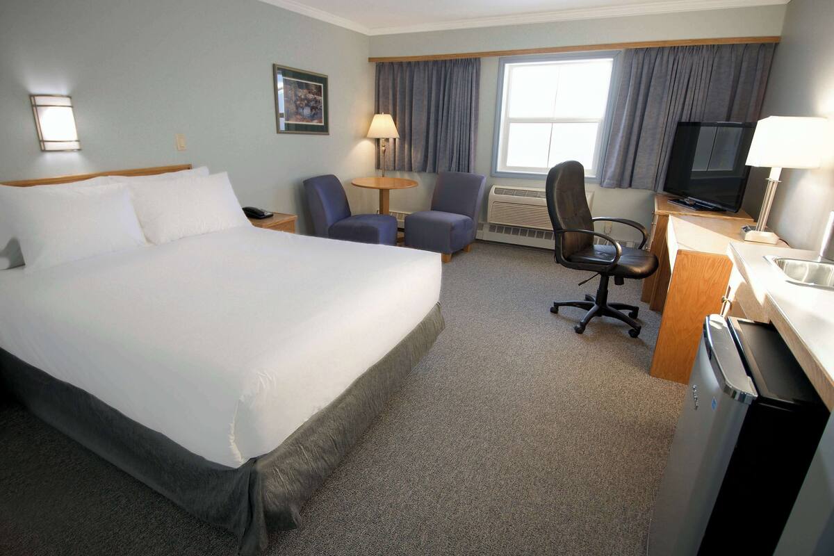 A hotel room with a large bed, two armchairs, a desk with a computer, a TV, and a window with curtains.