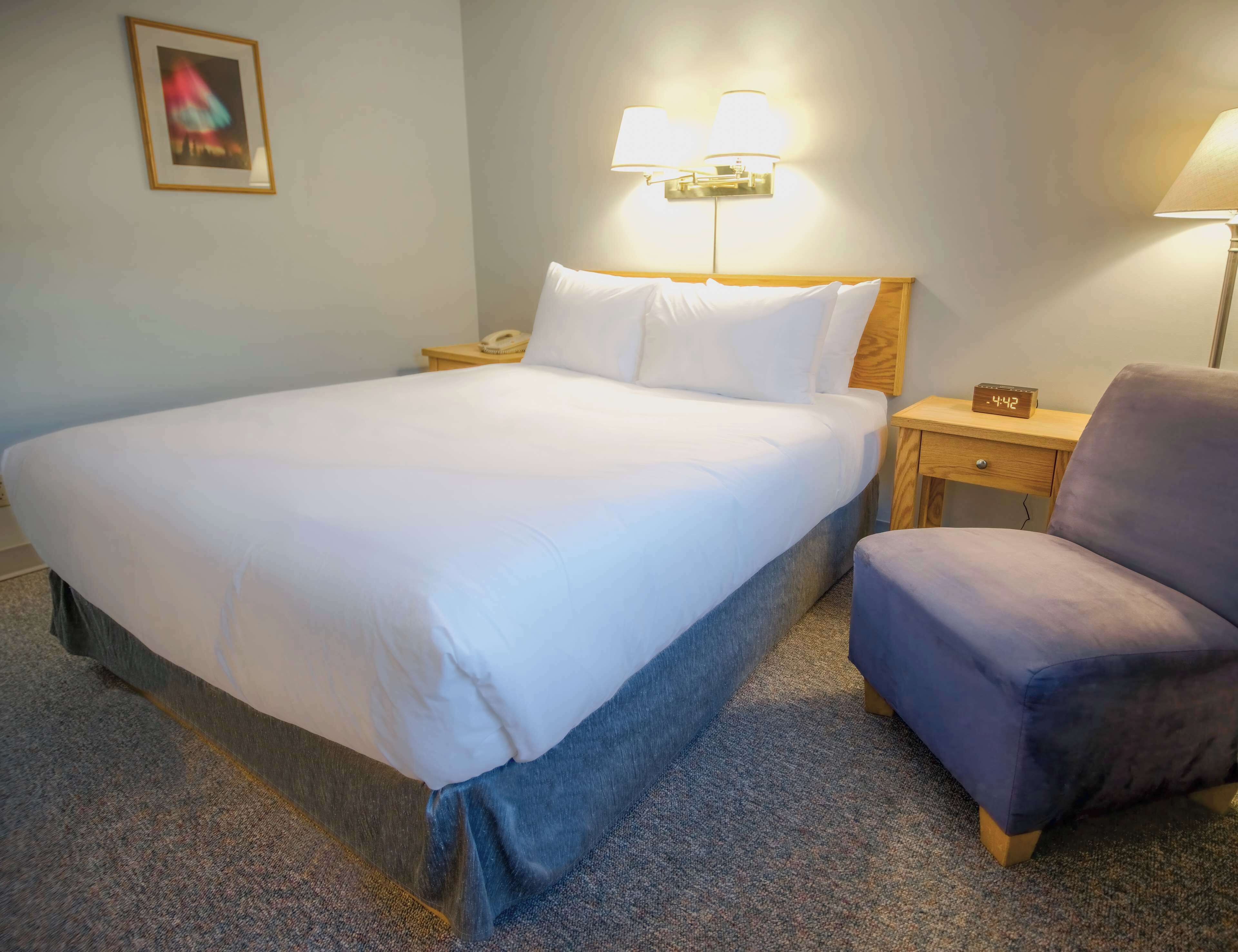 A hotel room with a large bed, a nightstand with a lamp, a chair, and a clock.