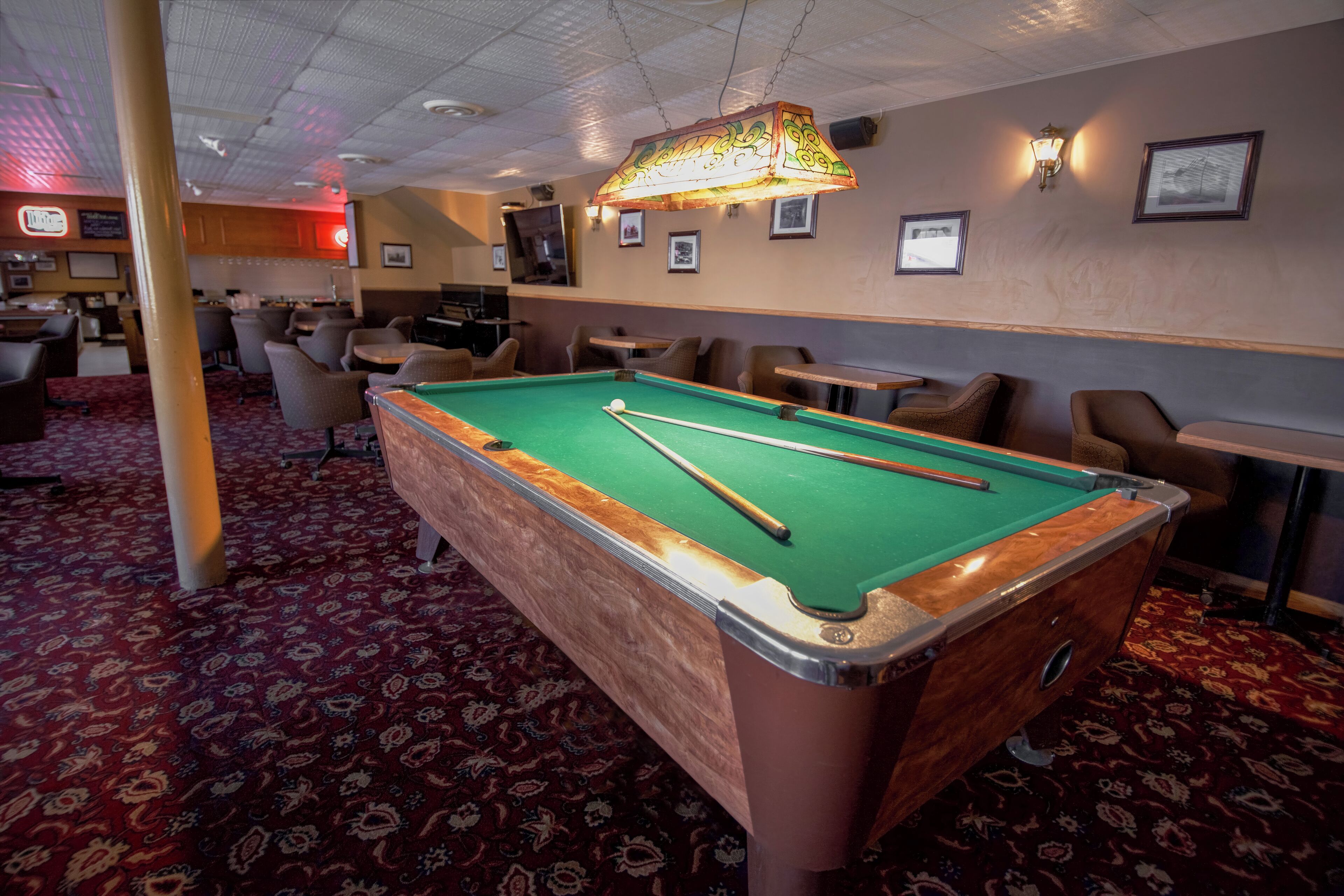 A pool table with a green felt surface, surrounded by chairs and tables in a room with patterned carpet.