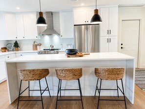 Une cuisine moderne avec des meubles blancs, un réfrigérateur en acier inoxydable, une cuisinière à gaz et trois tabourets de bar en osier.