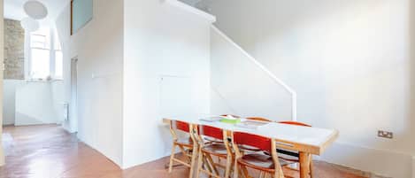 Une pièce avec un plancher en bois, une table blanche avec des chaises, un escalier et une fenêtre.