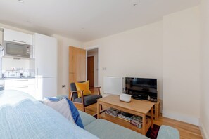 A modern living room with a sofa, a coffee table, a television, and a kitchen area with a microwave. - Mid Century 2 BDR Private Balcony Canary (London)