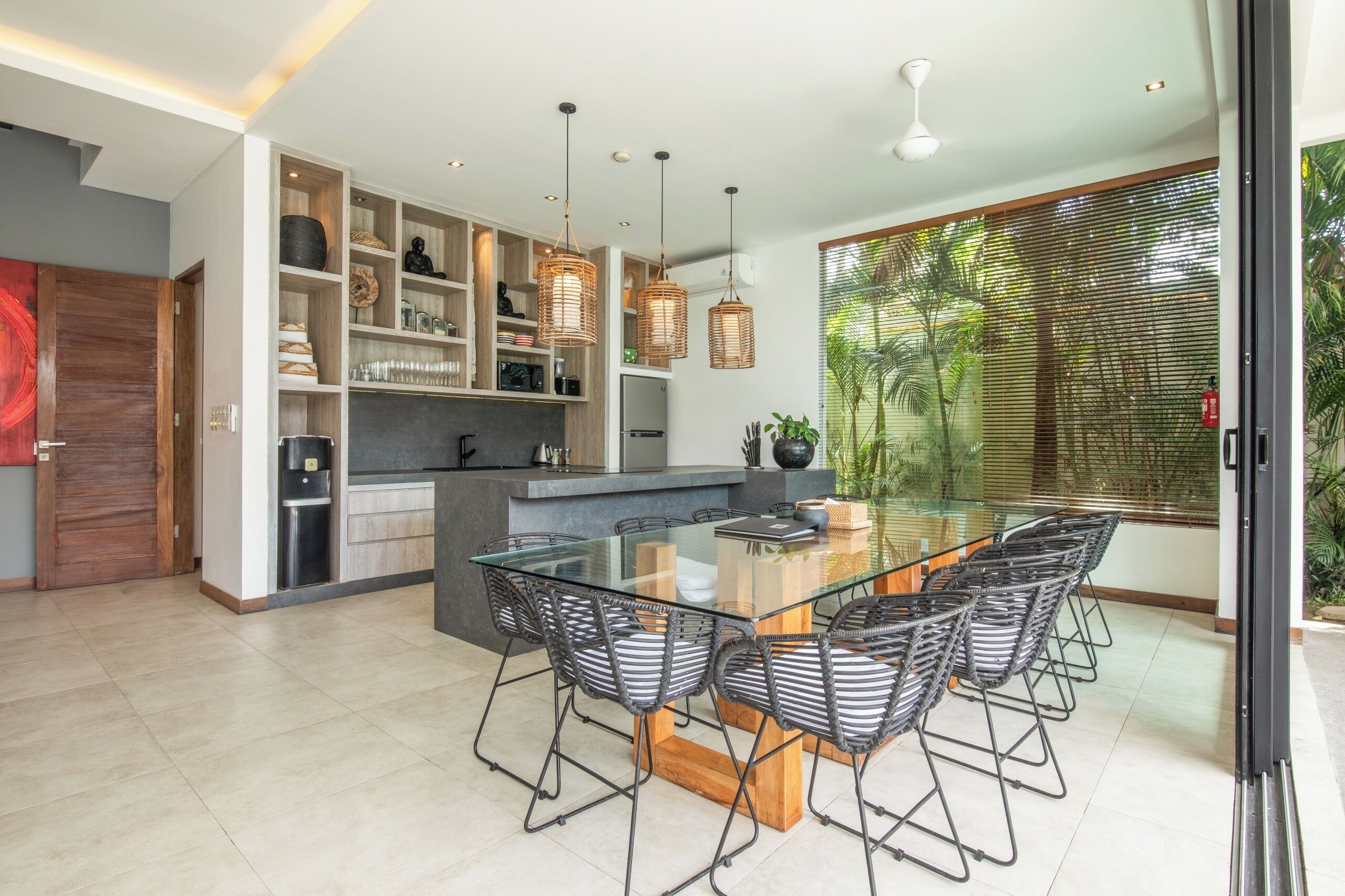 Une cuisine moderne avec une table à manger en verre, des suspensions en osier et une vue sur la verdure grâce à de grandes fenêtres.