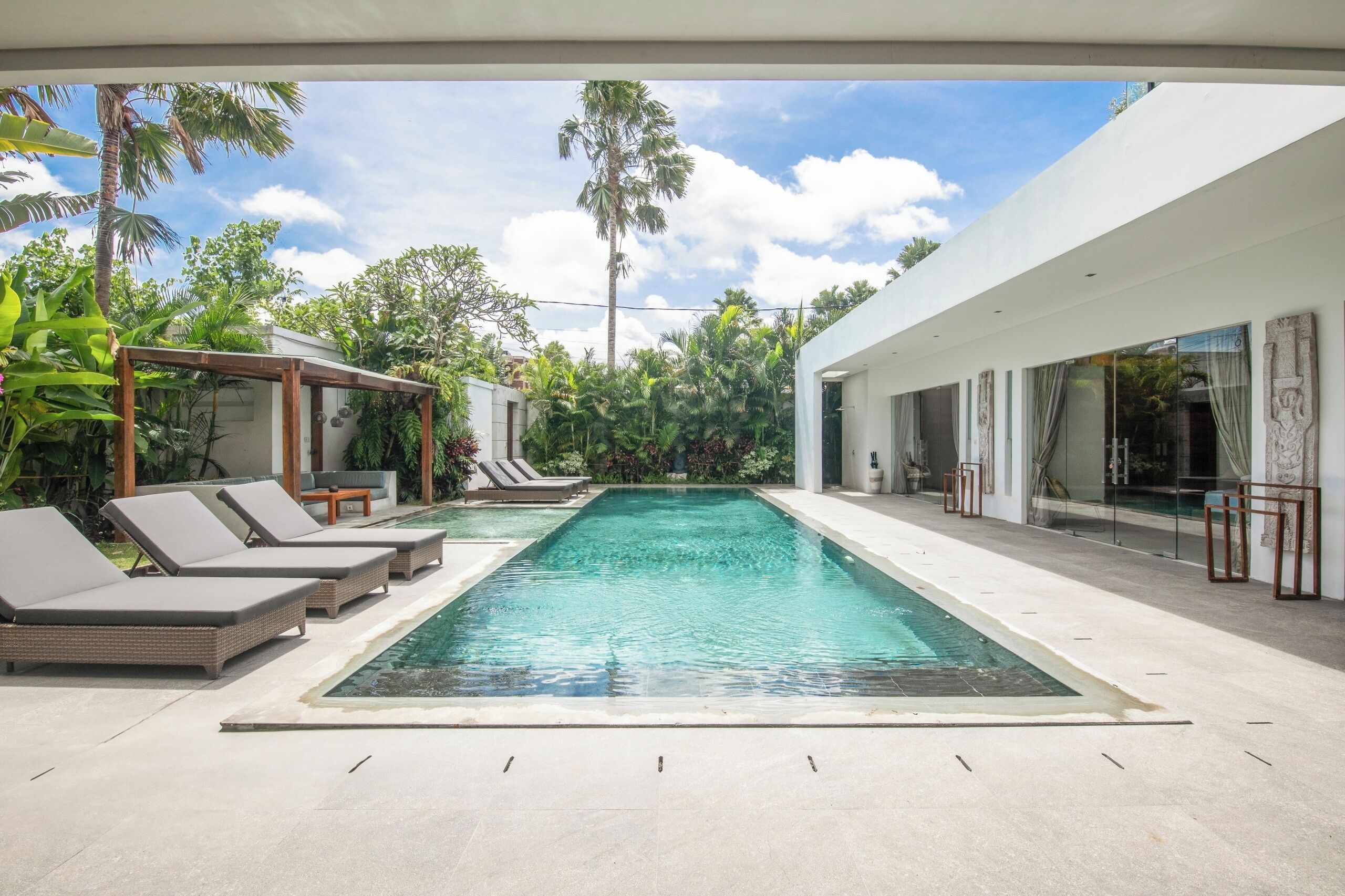 Un espace piscine moderne, doté de chaises longues et d’une piscine à eau claire, entouré d’une végétation luxuriante et d’un bâtiment blanc.