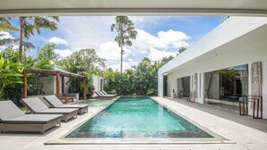 A modern pool area with lounge chairs and a clear pool, surrounded by lush greenery and a white building.