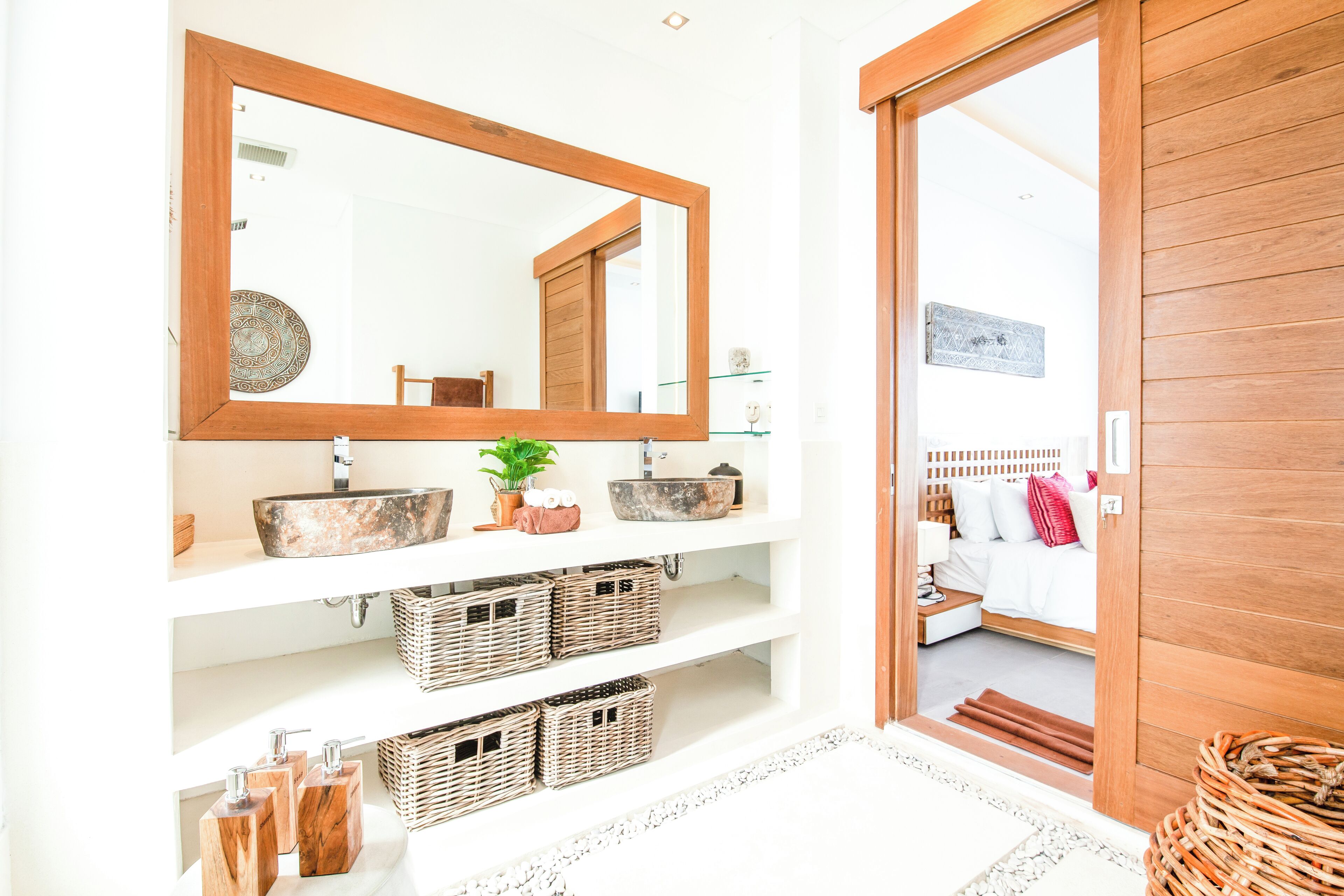 A bathroom with a large mirror, two sinks, wicker baskets, and a wooden door leading to a bedroom.