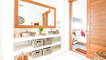 A bathroom with a large mirror, two sinks, wicker baskets, and a wooden door leading to a bedroom.