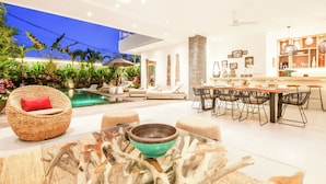 Un área de piscina interior moderna con zona de comedor, mobiliario de mimbre y una mesa de vidrio con un centro de mesa de madera.