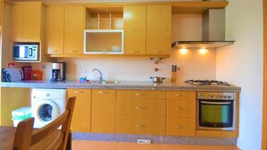 A compact kitchen with wooden cabinets, a microwave, a washing machine, a gas stove, and a built-in oven. - Estrela do Mar - Com Vista Para a Piscina (Alcobaça)