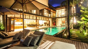 Zona junto a la piscina con sillones de descanso y sombrillas, una piscina y un edificio bien iluminado con espacios interiores a la vista.