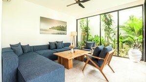 A modern living room with a sectional sofa, a wooden coffee table, and a wooden chair. There is a ceiling fan, a floor lamp, and a large window with plants visible through it. - New 4BR Villa 200m to the Beach Canggu (Munggu)