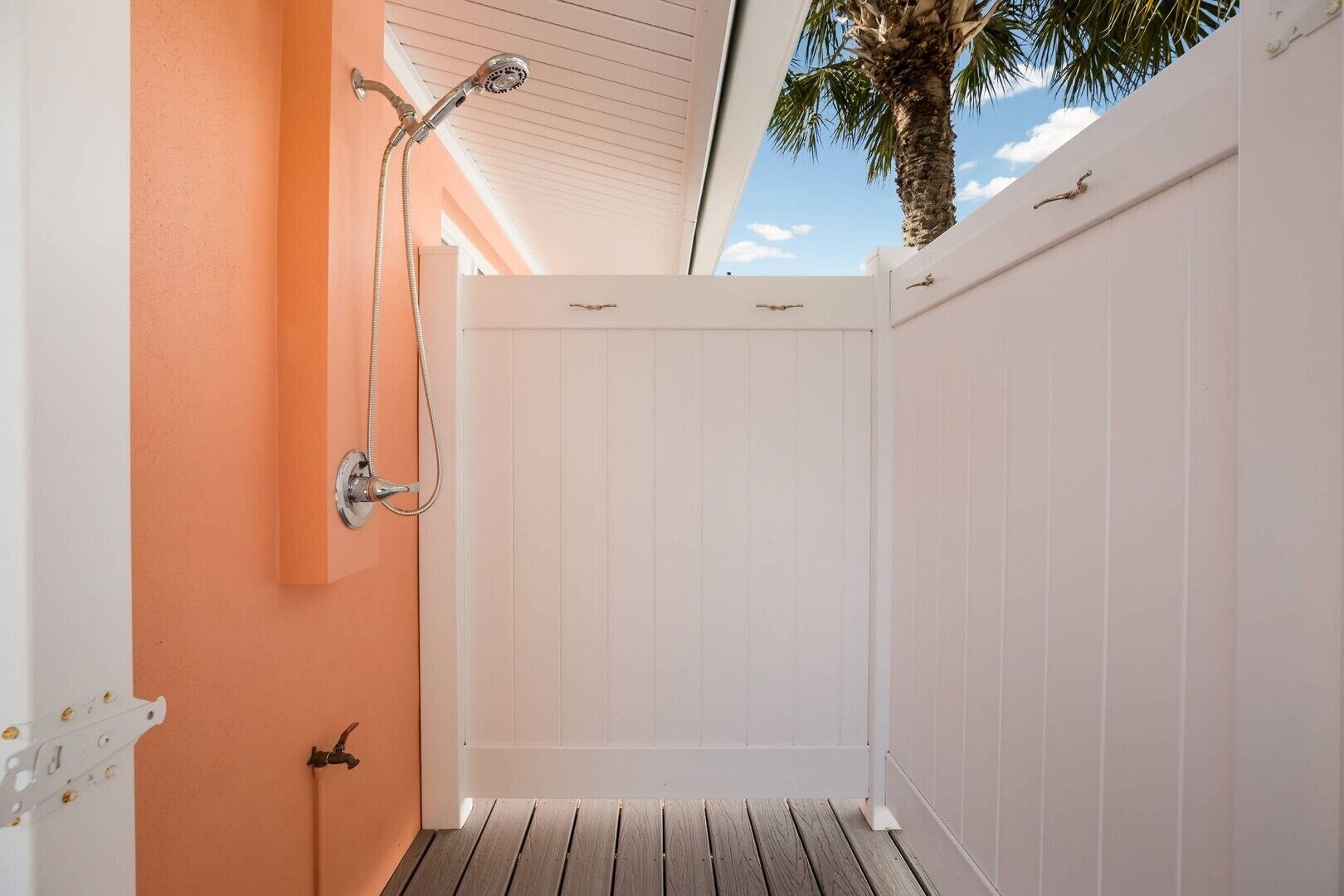 A covered outdoor shower area with a wooden deck, white walls, and a palm tree in the background.