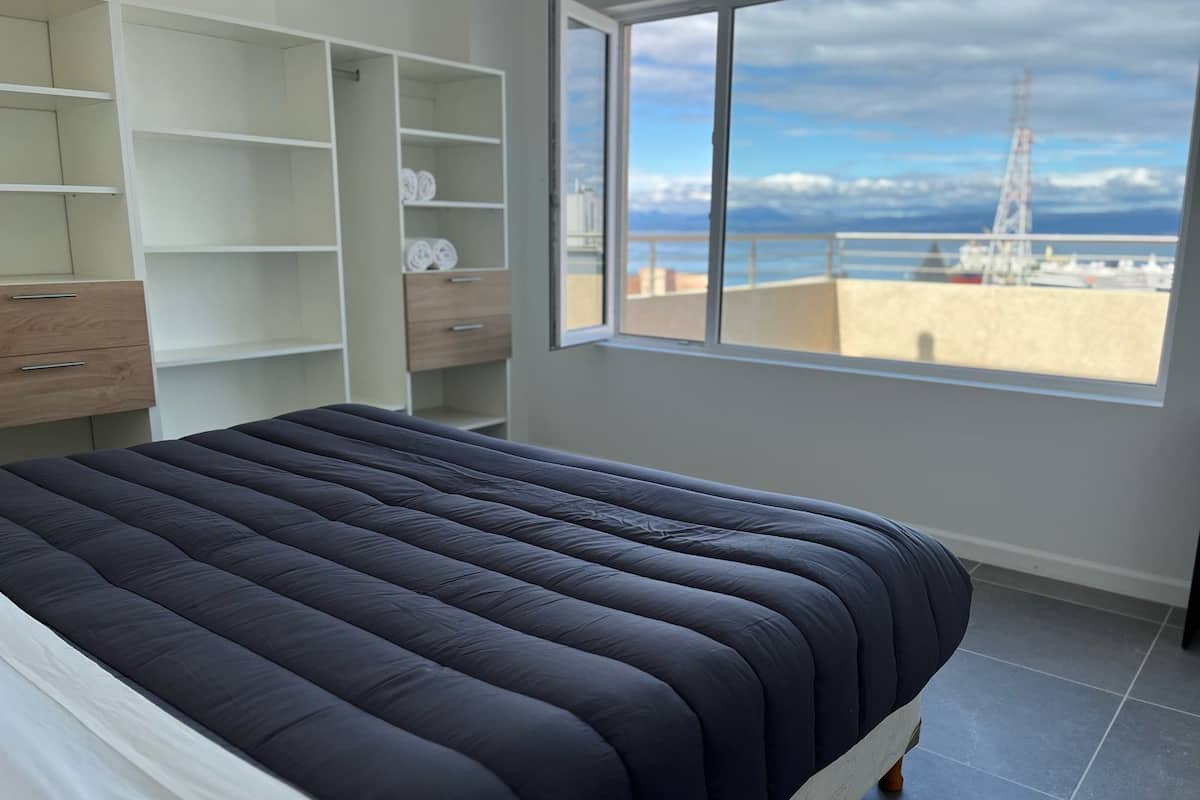 A bedroom with a large bed, a wardrobe, and a window with a view of the sea and mountains.
